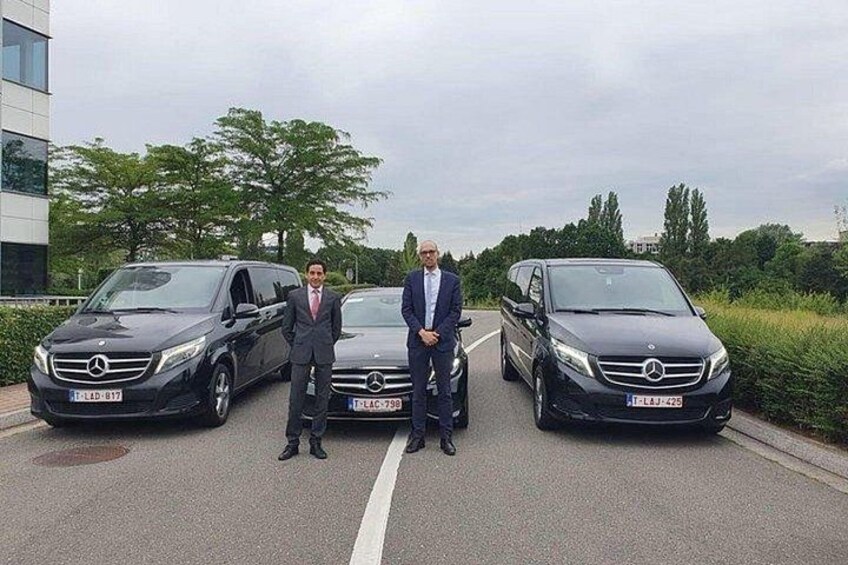 Two men standing between two black Mercedes-Benz vehicles on a road with trees and a building in the background. 2