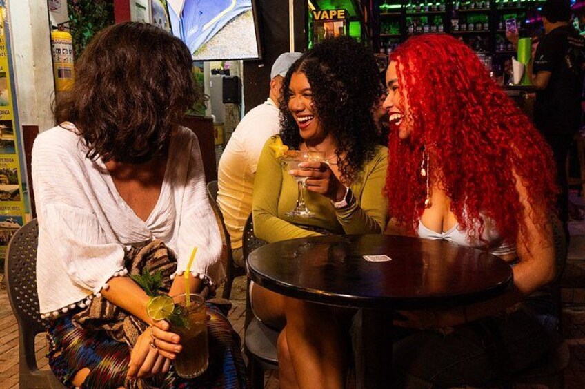 Trois personnes sont assises à une table, devant des boissons. On aperçoit un bar en arrière-plan et une enseigne sur laquelle on peut lire « VAPE ». 2