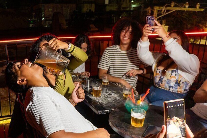Un grupo de personas sentadas en un bar, algunas sosteniendo bebidas, y alguien usando un teléfono inteligente. 3