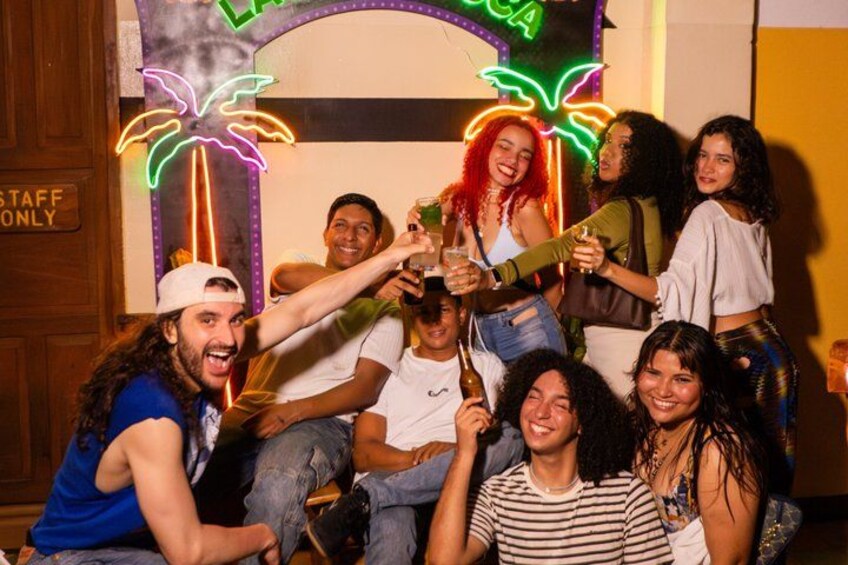 A group of people toasting with drinks in a room with neon palm tree decorations and a 'STAFF ONLY' sign. 3