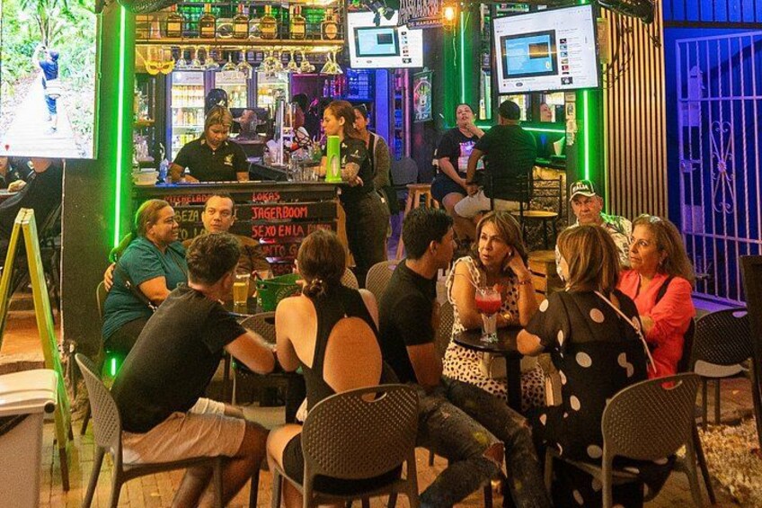A bar with people seated at tables, a bartender, and a counter with drinks and a menu. 2
