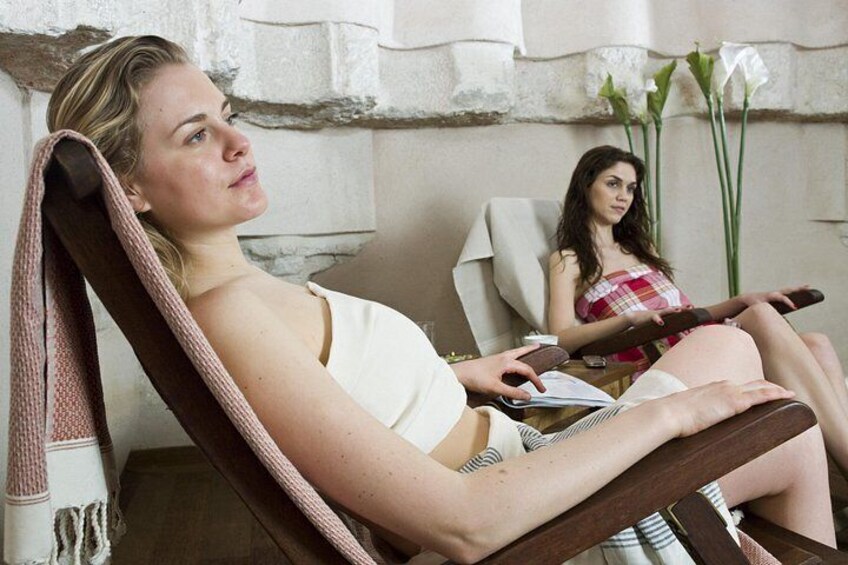 Two women sitting in a room with a stone wall and a window with white flowers. 3