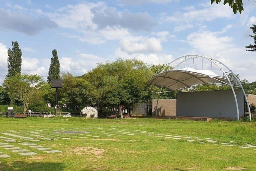Ein Park mit einem weißen Kiosk, einer Steinskulptur und einer Rasenfläche mit Steinwegen. 2