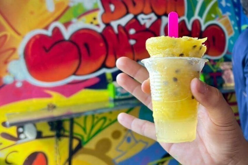 Une main tenant un gobelet en plastique contenant une boisson jaune, avec une tranche d’ananas posée sur le dessus, sur fond d’un mur recouvert de graffitis colorés. 4