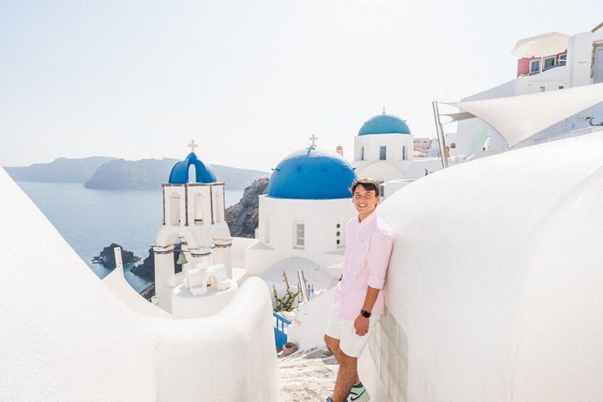 A person in a pink shirt and white shorts standing on a white building with blue domes, overlooking a coastal landscape with white buildings and blue domes. 4