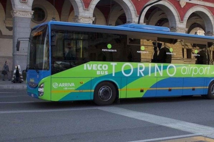 A blue and green bus with 'Torino airport' and 'Arriva' branding, parked in front of a building with arches. 5