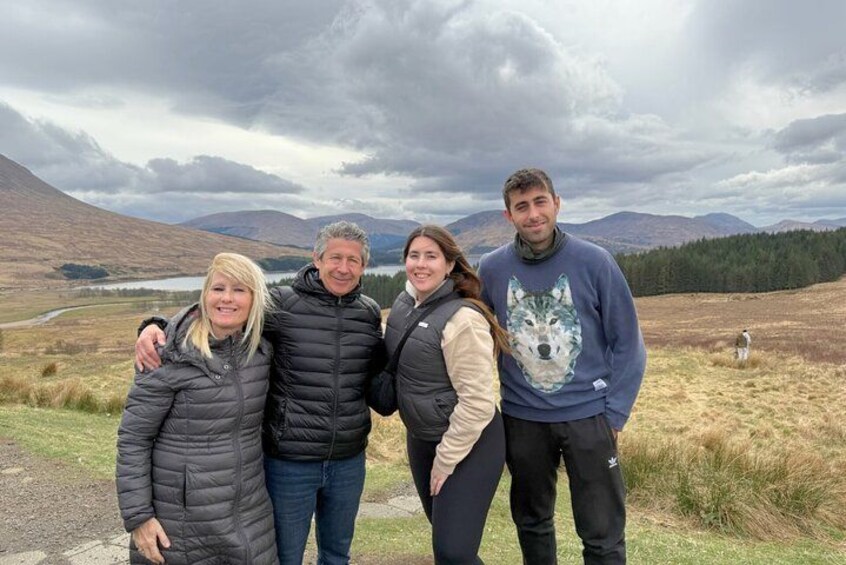 Four people standing on a path with a mountainous landscape and a lake in the background. 3