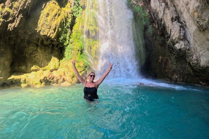 A person in a black swimsuit standing in clear blue water with a waterfall in the background. 1