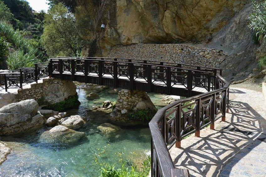 A wooden bridge over a clear stream with rocky banks, surrounded by greenery and a stone wall. 2