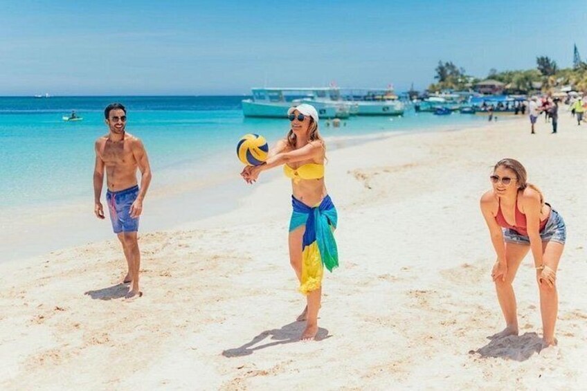 Una playa con gente jugando voleibol, un barco al fondo y cielos despejados. 3