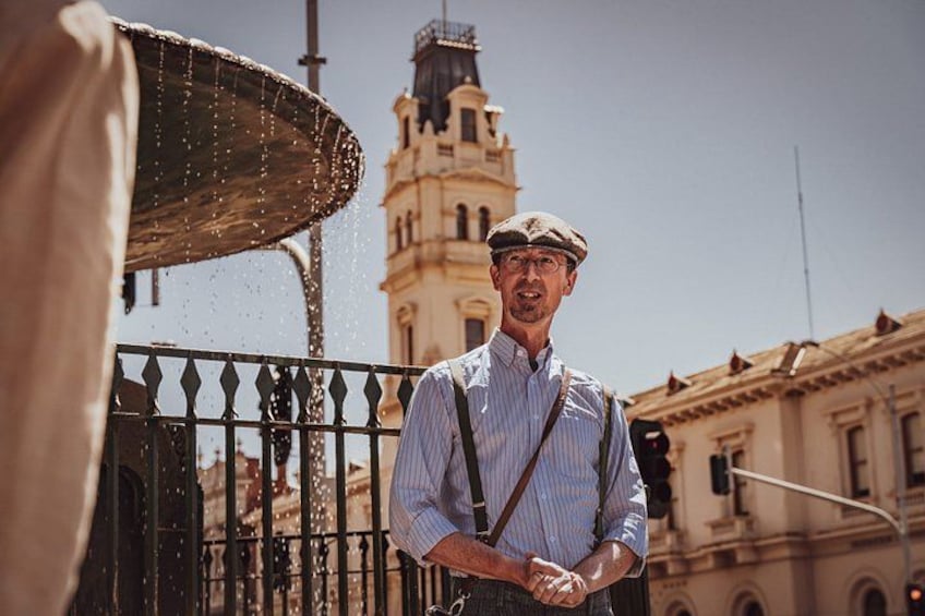 A person standing in front of a building with a tower, wearing a hat and suspenders. 1
