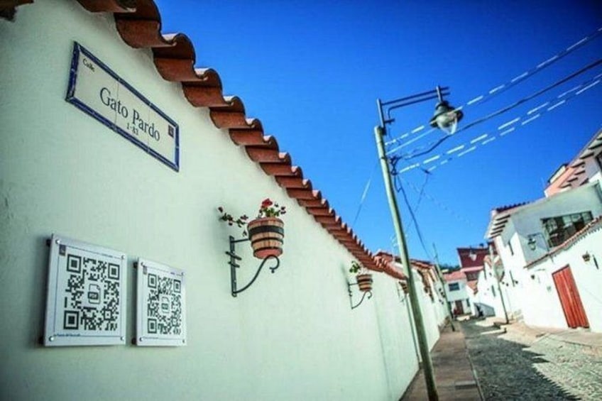 A narrow street with a white building labeled 'Gato Pardo,' a hanging flower pot, and two QR codes on the wall. 1