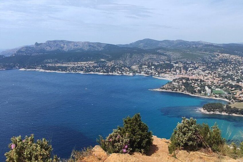 A coastal town with a clear blue sea, a mountain range in the background, and sparse vegetation in the foreground. 1
