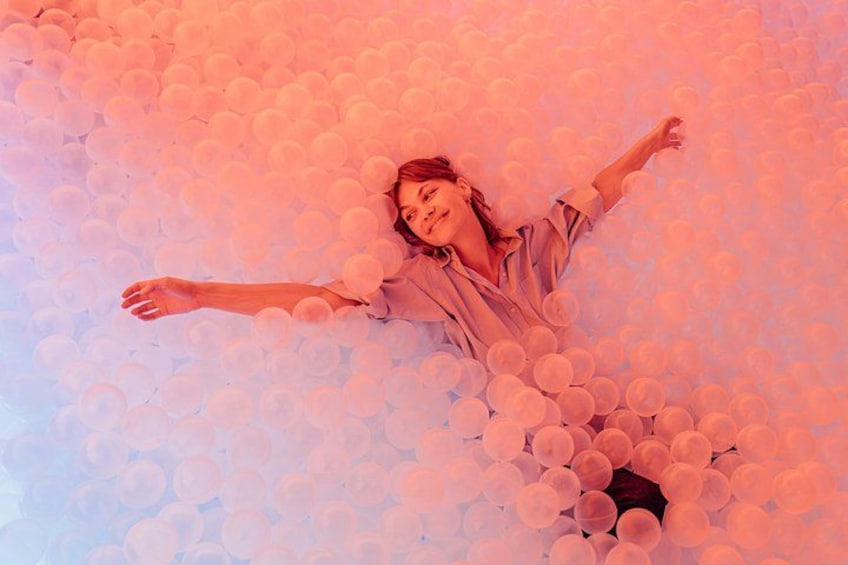 Una persona rodeada de numerosas esferas blancas, posiblemente globos, en una habitación de tono rosado. 1
