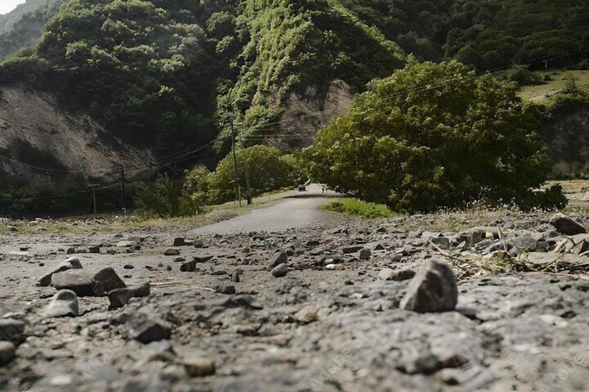 A rocky path with a road in the distance, surrounded by greenery and hills. 1