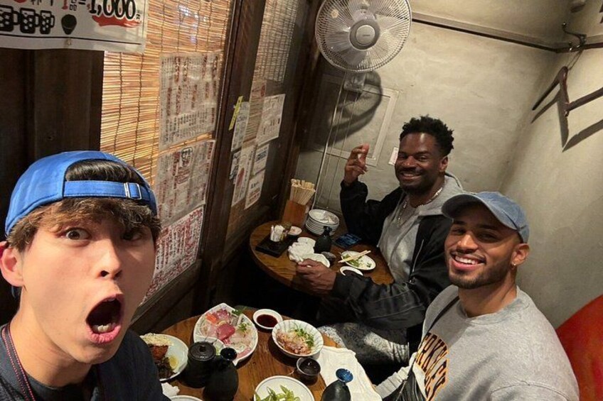 Three people sitting at a table with food and drinks, in a room with a fan and papers on the wall. 2