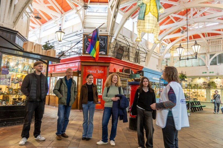 A group of people standing in a covered market area with shops and a red kiosk in the background. 2