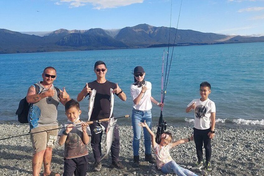 Un grupo de personas junto a un lago, con cañas de pescar, cielo despejado y montañas al fondo. 1