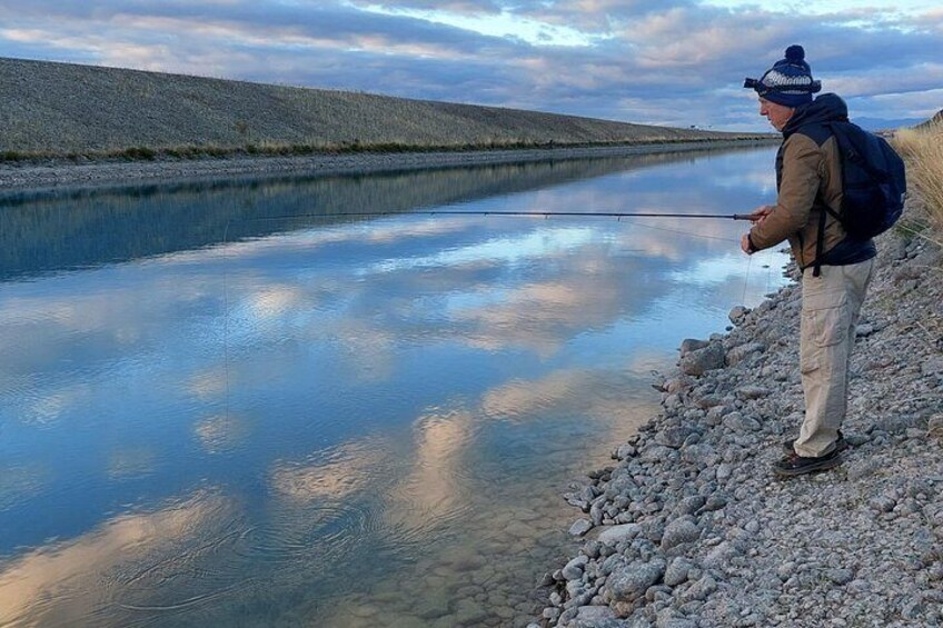 A person fishing by a calm lake with a rocky shoreline and distant hills. 5