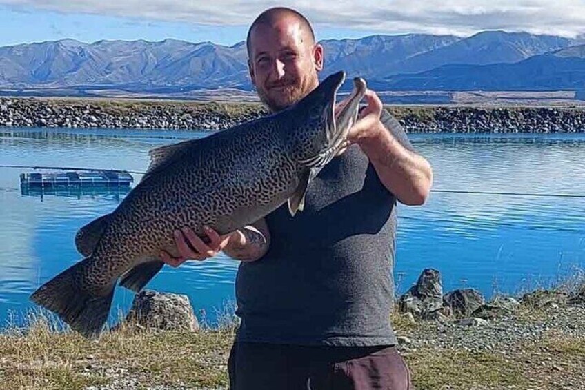 A person holding a large fish by a lake with mountains in the background. 1