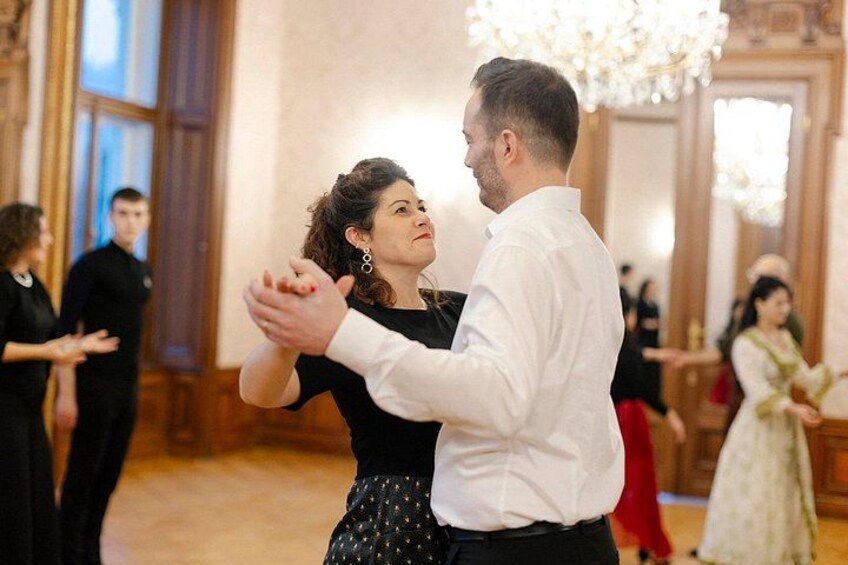 A couple dancing in a ballroom with chandeliers and other guests in the background. 1
