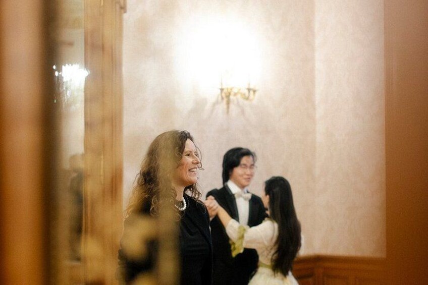 A person in a black suit with a white shirt and tie, standing in a room with a chandelier. 2
