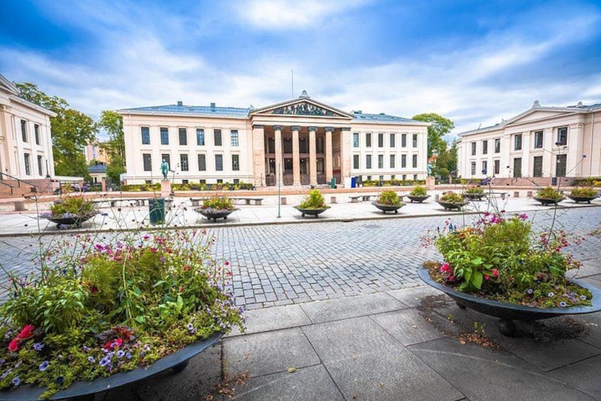En klassisk byggnad med kolonner, omgiven av blomsterrabatter och ett stenlagt torg. 5