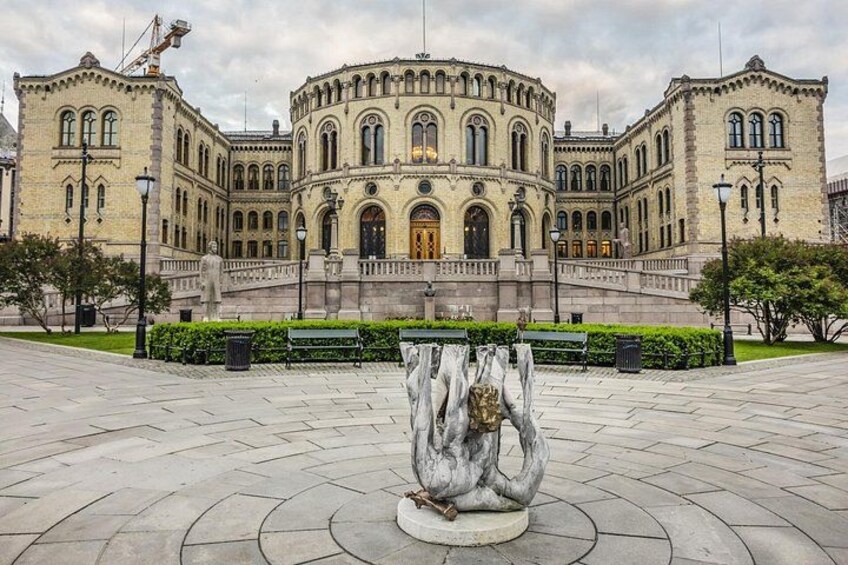 En historisk byggnad med välvda fönster och en central entré, omgiven av ett stenbelagt torg med en skulptural installation. 3