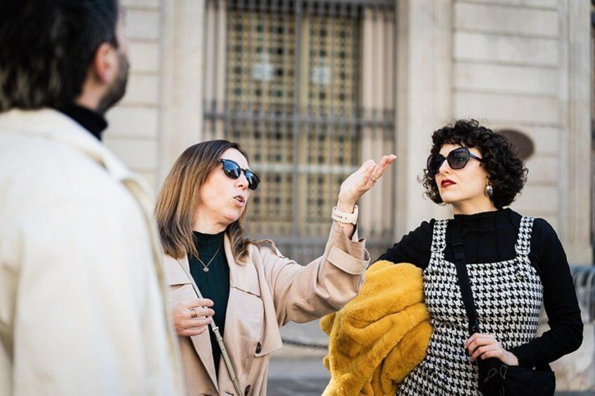 Two people in conversation, one wearing a beige coat and the other in a black and white patterned dress with a yellow scarf. 5