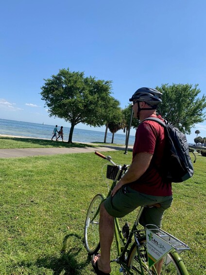 A person on a bicycle with a green frame, wearing a helmet and a red shirt, parked on grass near a body of water with trees and a clear sky. 1