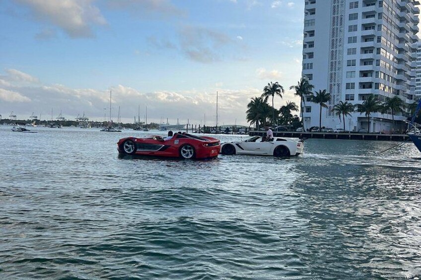 Two cars partially submerged in water near a marina with a tall building and palm trees in the background. 3