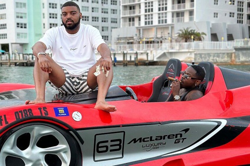 A red McLaren GT car with two people on it, near a waterfront with buildings in the background. 5