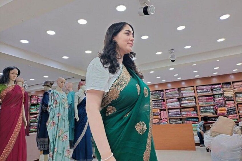 Eine Frau in einem grünen Saree in einem Geschäft mit Stoffregalen und Kleiderträgerpuppen. 1