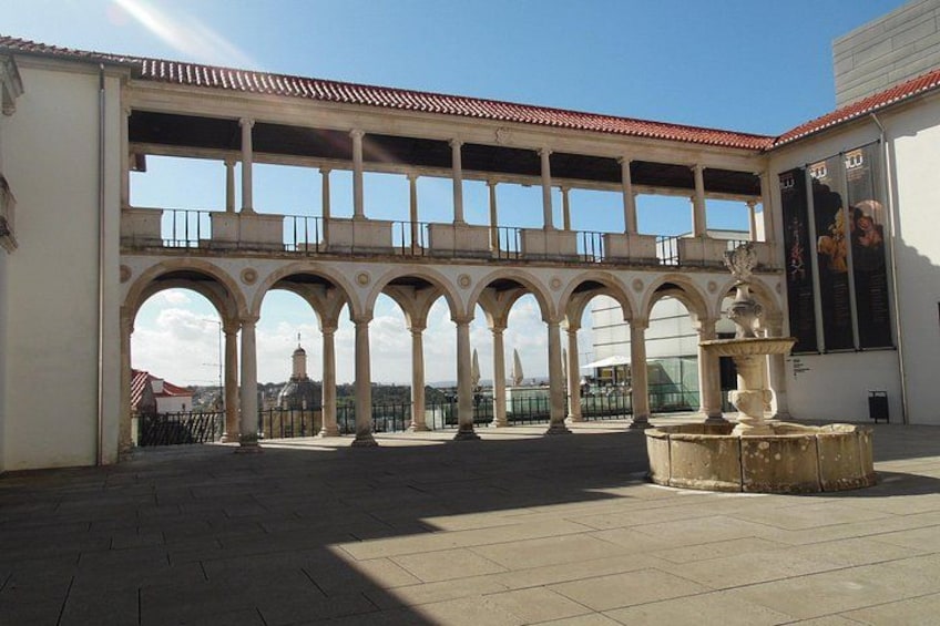 A courtyard with arches, a fountain, and a building with a red roof. 1