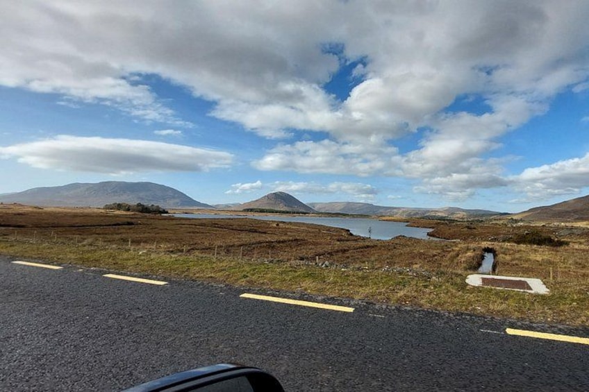 A road with yellow lines, a grassy field, a small body of water, and distant mountains under a partly cloudy sky. 1