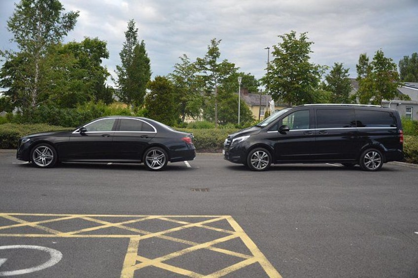 A black sedan and a black van parked in a parking lot with yellow parking lines and trees in the background. 3