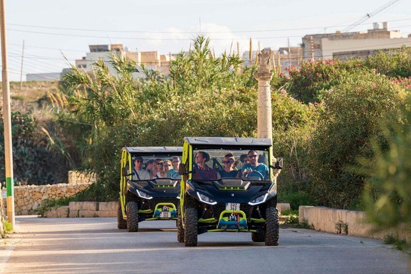 Two off-road vehicles on a paved road with greenery and buildings in the background. 1