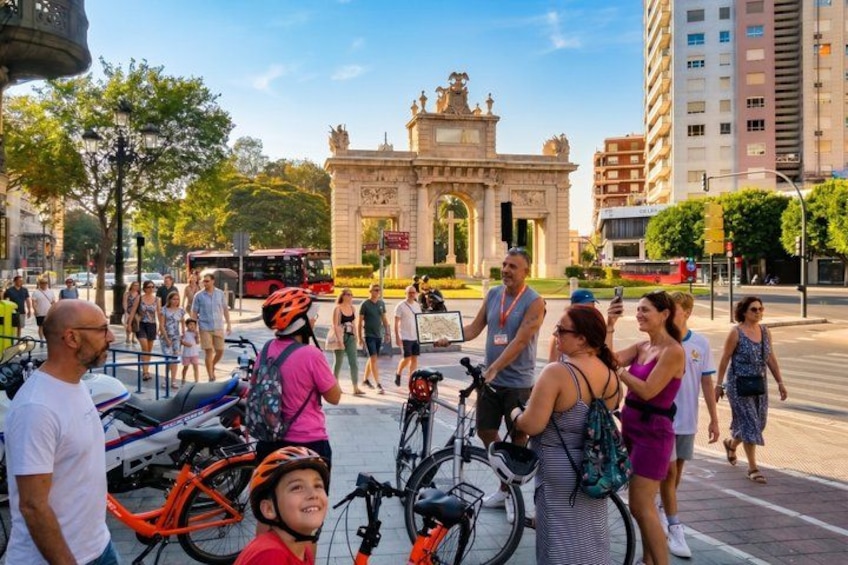 Una calle urbana concurrida, con gente, bicicletas y un arco histórico al fondo. 4
