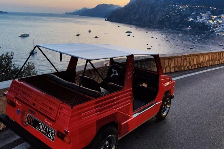 A red vintage car parked on a coastal road with a scenic view of the sea and boats. 1