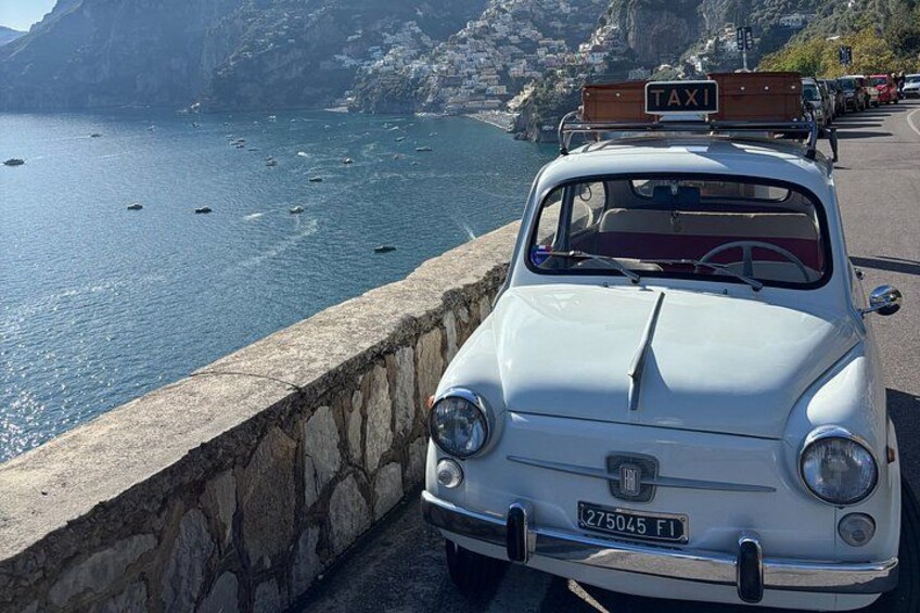 A white vintage car with a 'TAXI' sign on top, parked by a stone wall overlooking a coastal area with boats and cliffs. 3