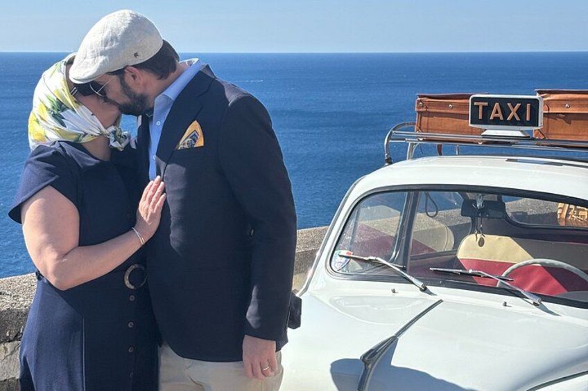 A couple standing by a white car with a 'TAXI' sign, a blue ocean in the background, and a clear sky. 4