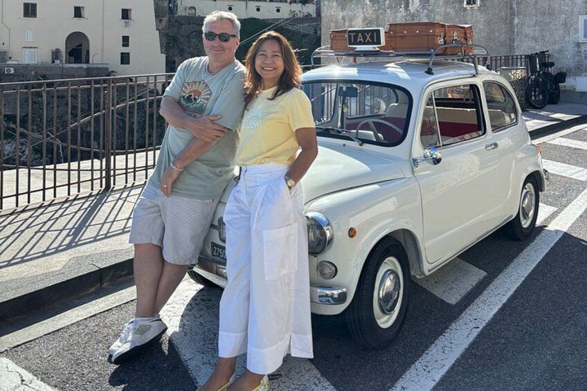 A vintage white car with a 'TAXI' sign on top, parked on a street with a metal railing and buildings in the background. 2