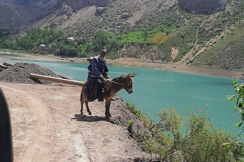 Una persona cavalca un asino lungo un sentiero sterrato accanto a un fiume, con montagne sullo sfondo. 2