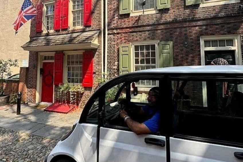A vintage car parked in front of a brick building with red shutters and an American flag. 2
