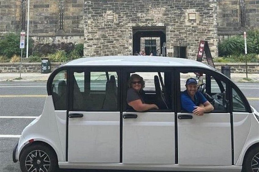 A white car with two people inside, parked on a street with a stone building in the background. 3