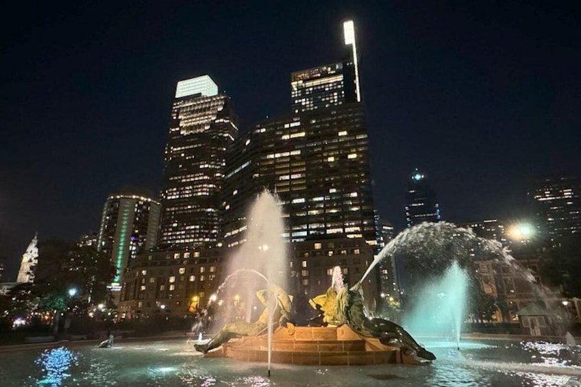 A cityscape at night with illuminated skyscrapers and a fountain with water jets. 3