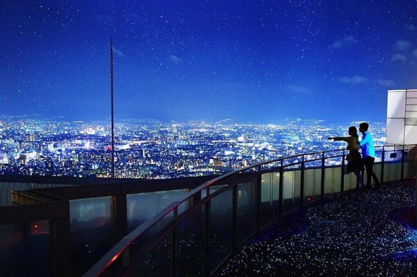 A night view of a cityscape with a starry sky, a tall flagpole, and two people standing on a balcony. 2