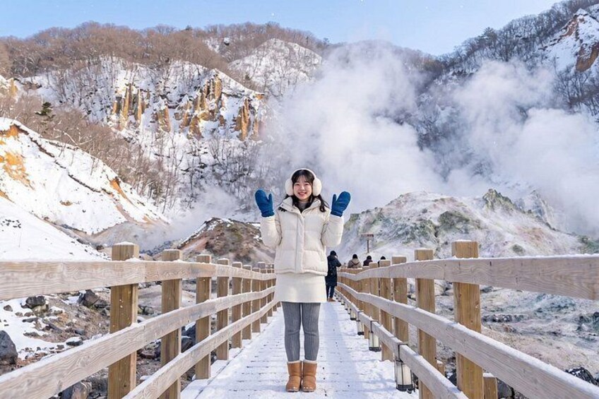 Una persona de pie sobre un puente de madera, con colinas nevadas y vapor elevándose al fondo. 1