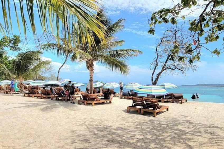 A beach with wooden lounge chairs and striped umbrellas, palm trees, and a clear blue sky. 1