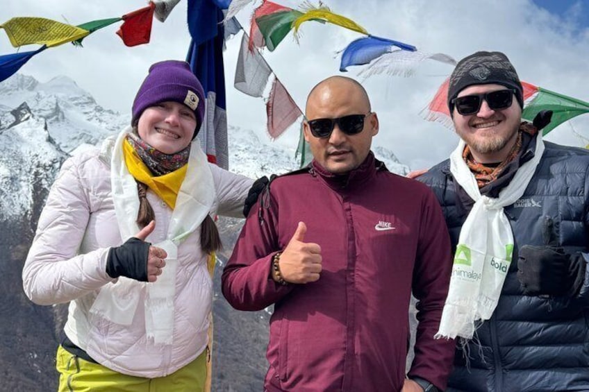 Three people standing with thumbs up, in front of a snowy mountain landscape with colorful prayer flags. 4